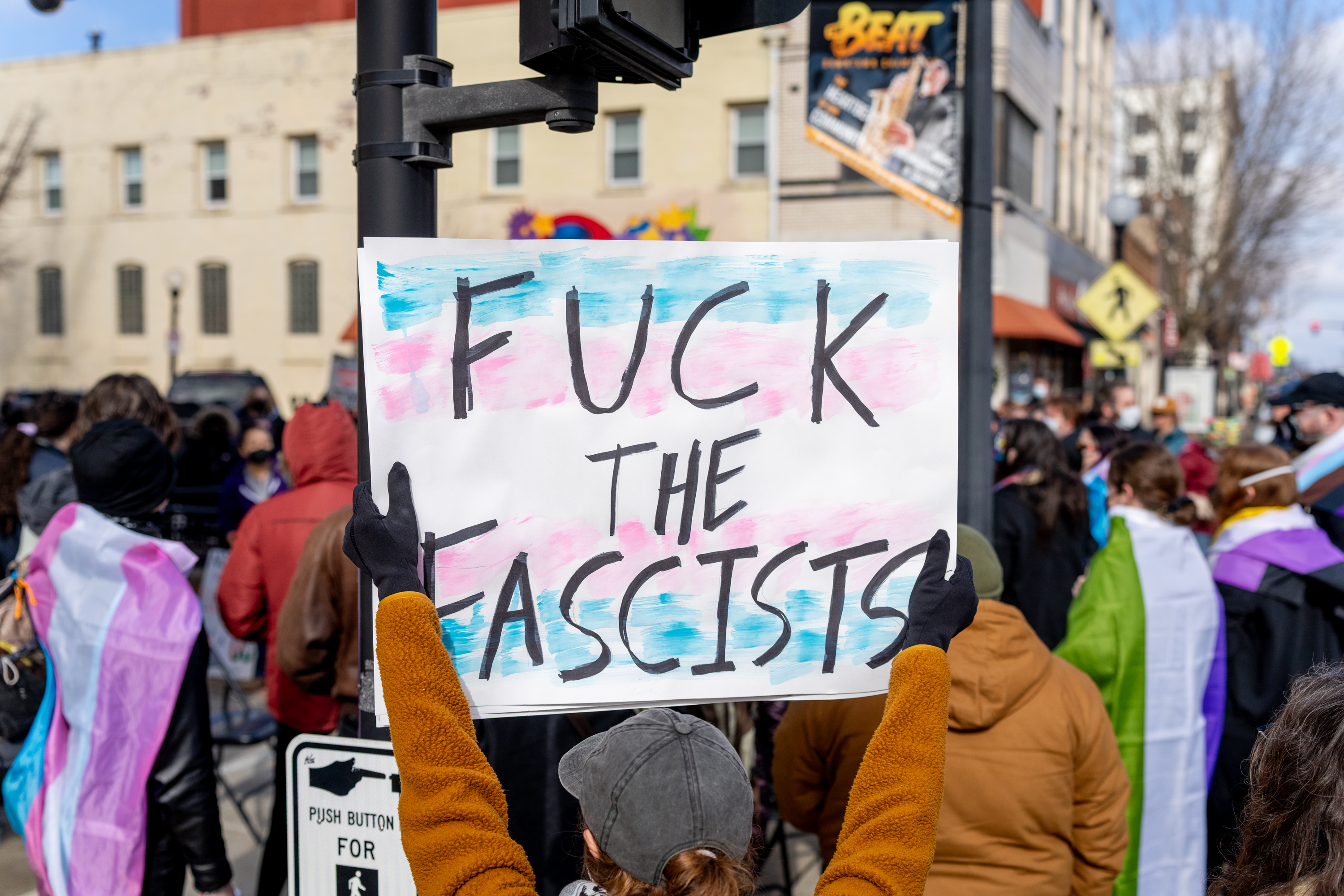 A photo of me (from behind) in a crowd holding a protest sign featuring a trans pride flag background with the words `Fuck the fascists'.